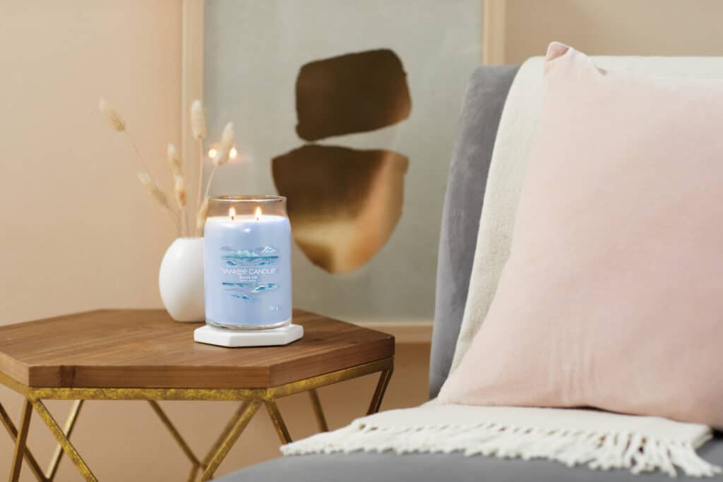 Blått doftljus på träbord bredvid rosa kudde och konstverk.