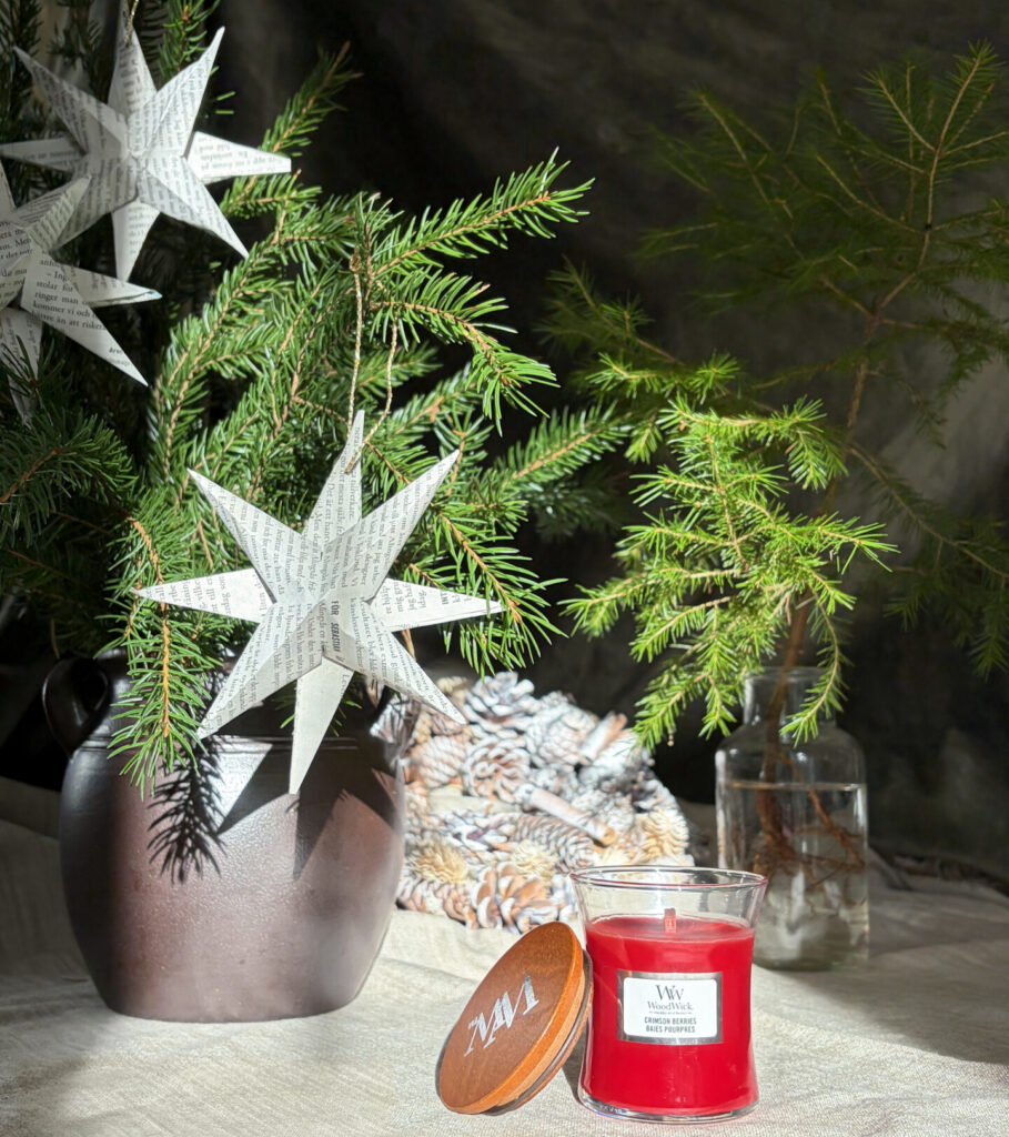 Juldekoration med granris, pappersstjärnor och röd doftljus på beige duk.