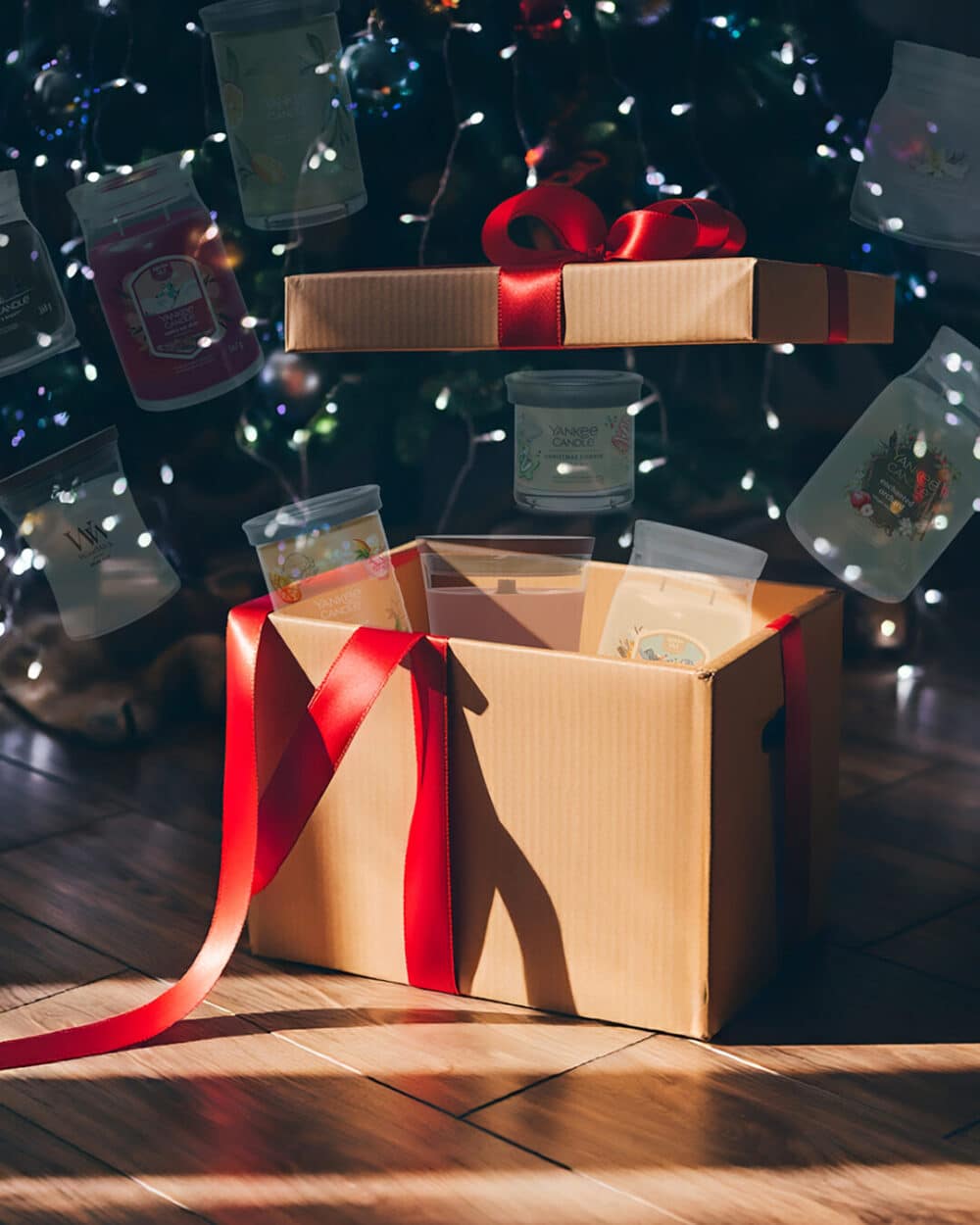 Presentask med rött band, fylld med doftljus framför dekorerad gran.