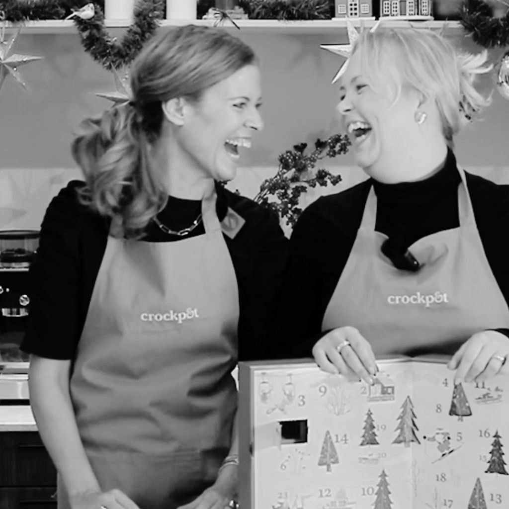 Två glada personer i köket, skrattande iklädda Crock-Pot förkläden under julförberedelser.