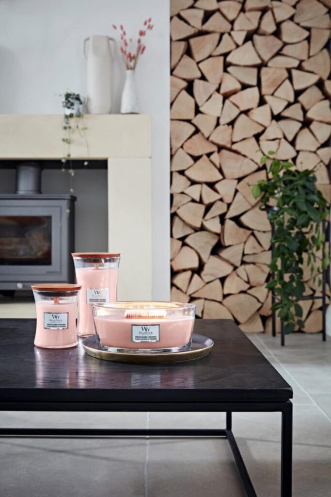 Rosa WoodWick-ljus på bord vid mysig braskamin och vedförråd.